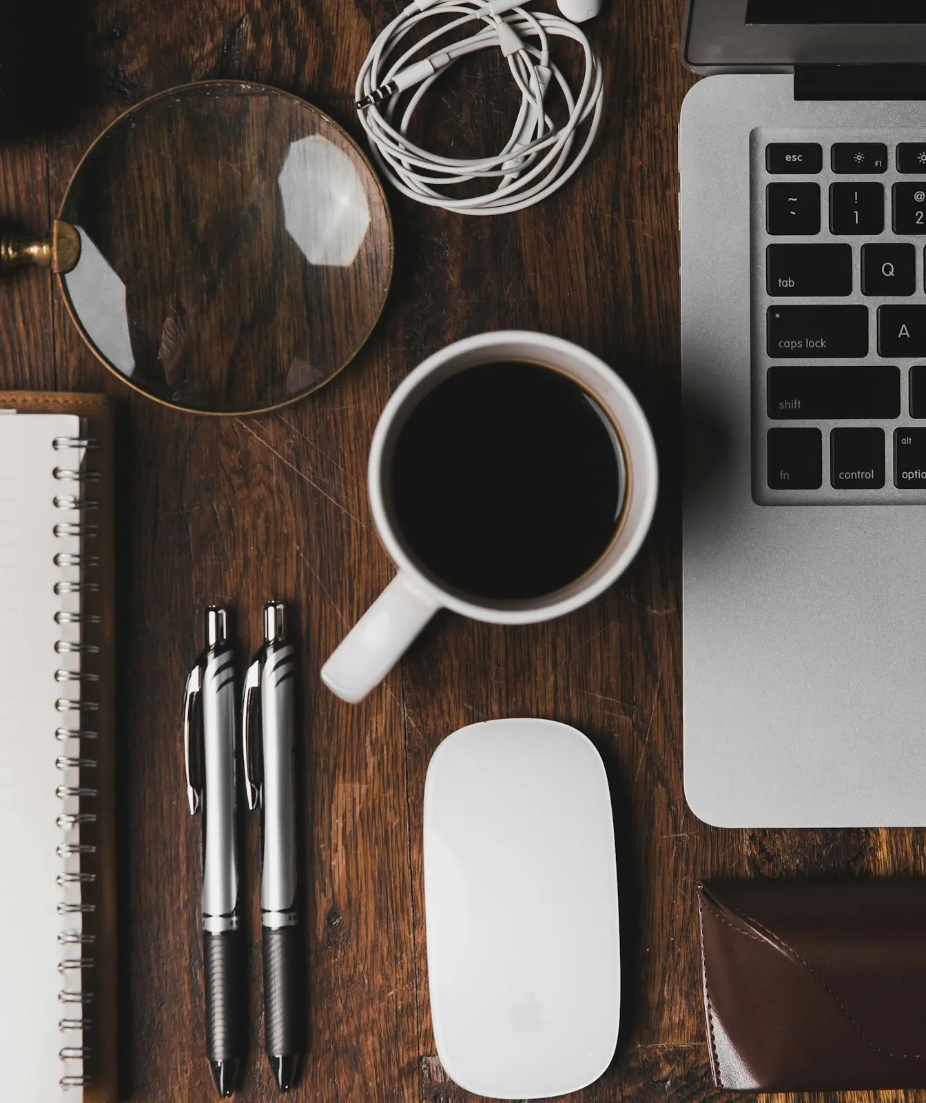 A laptop, notebook, pens and a cup of coffee on a wooden desk, seen from above